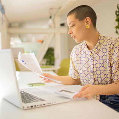 Young woman holding calculator while looking at forms at a desk with a laptop on it.