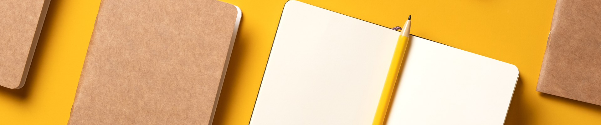 Four notebooks arranged on a yellow desk, one with a pencil on it.